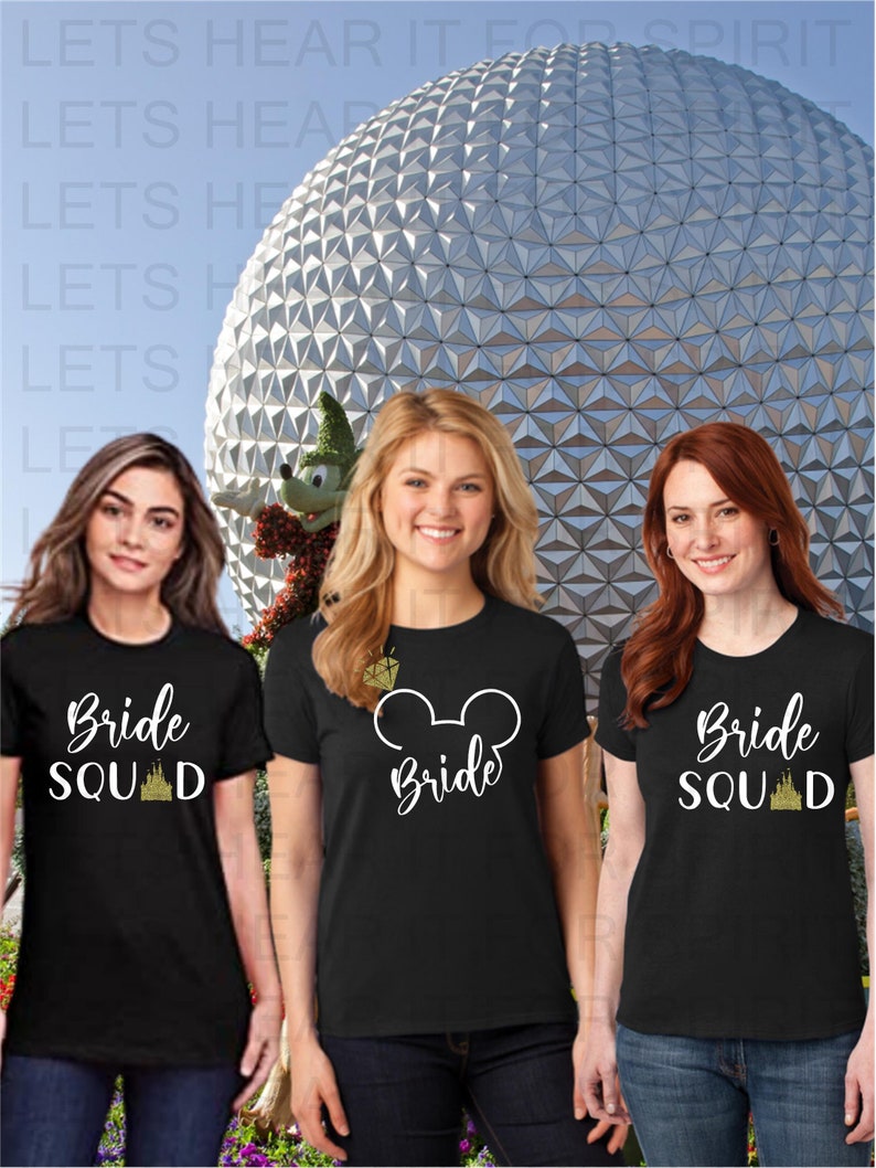 May include: Three women wearing black t-shirts with white text and gold glitter accents. The shirts say "Bride Squad" and "Bride" with a Mickey Mouse ear graphic. The women are standing in front of a large silver sphere.