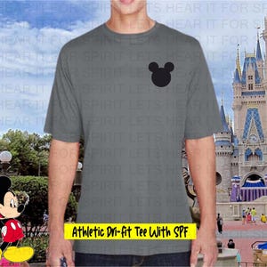 May include: A gray athletic t-shirt with a black Mickey Mouse head graphic. The text "Athletic Dri-fit Tee With SPF" is displayed in a yellow rectangle. The background features a theme park with a castle and cartoon character.