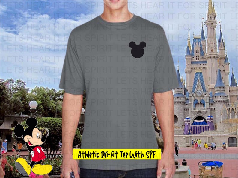 May include: A gray athletic t-shirt with a black Mickey Mouse head graphic. The text "Athletic Dri-fit Tee With SPF" is displayed in a yellow rectangle. The background features a theme park with a castle and cartoon character.