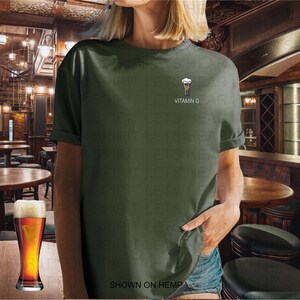 K&ouml;nnte beinhalten: Olivgr&uuml;nes T-Shirt mit einer kleinen Grafik eines Bierglases und dem Text "VITAMIN G". Ein volles Bierglas steht auf einem Holztisch vor der Person, die das T-Shirt tr&auml;gt. Der Text "SHOWN ON HEMP" steht unten.