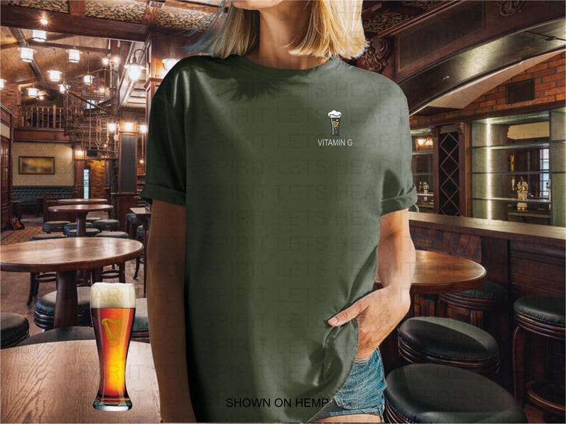 K&ouml;nnte beinhalten: Olivgr&uuml;nes T-Shirt mit einer kleinen Grafik eines Bierglases und dem Text "VITAMIN G". Ein volles Bierglas steht auf einem Holztisch vor der Person, die das T-Shirt tr&auml;gt. Der Text "SHOWN ON HEMP" steht unten.