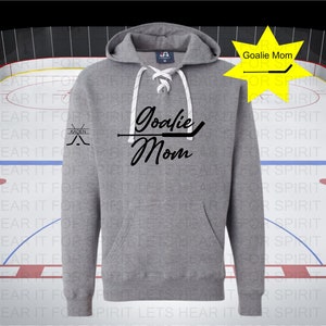 K&ouml;nnte beinhalten: Grauer Hoodie mit einem wei&szlig;en Kordelzug und einem schwarzen Aufdruck, der "Goalie Mom" mit einem Hockeyschl&auml;ger &uuml;ber dem Text sagt. Der linke &Auml;rmel hat ein kleines schwarzes Motiv aus zwei gekreuzten Hockeyschl&auml;gern mit dem Wort "Kaden" dar&uuml;ber.