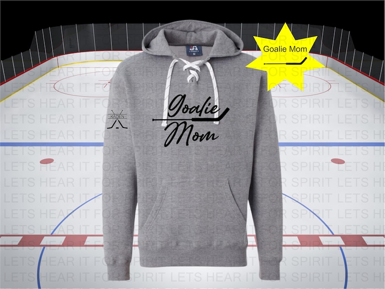 K&ouml;nnte beinhalten: Grauer Hoodie mit einem wei&szlig;en Kordelzug und einem schwarzen Aufdruck, der "Goalie Mom" mit einem Hockeyschl&auml;ger &uuml;ber dem Text sagt. Der linke &Auml;rmel hat ein kleines schwarzes Motiv aus zwei gekreuzten Hockeyschl&auml;gern mit dem Wort "Kaden" dar&uuml;ber.