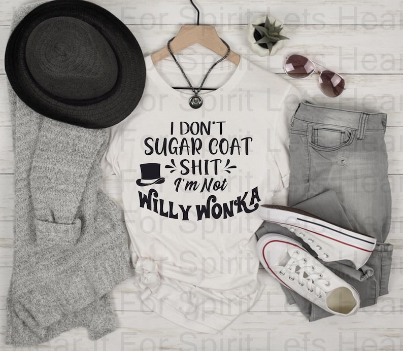 May include: A white t-shirt with the black text "I don't sugar coat shit, I'm not Willy Wonka". The shirt is styled with grey jeans, white trainers, a black hat, and a grey cardigan. Sunglasses and a necklace are also present.
