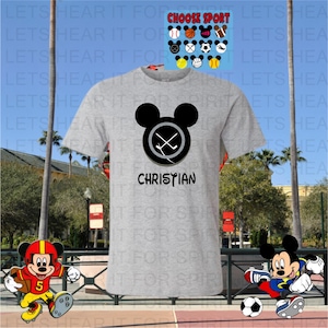 May include: A light gray t-shirt featuring a black Mickey Mouse head with crossed hockey sticks inside a circle. The name "CHRISTIAN" is printed below. Additional sports-themed Mickey Mouse designs are displayed above.