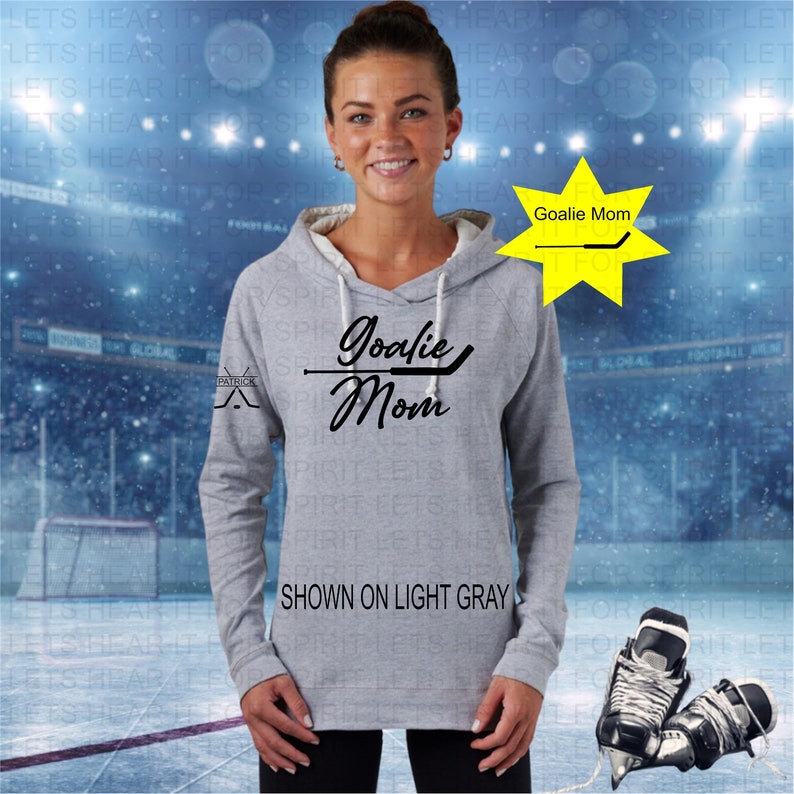Puede incluir: Una sudadera con capucha gris con el texto "Goalie Mom" y un gr&aacute;fico de un palo de hockey. El texto "Goalie Mom" tambi&eacute;n se muestra en una estrella amarilla en la esquina superior derecha de la imagen. El texto "SHOWN ON LIGHT GRAY" est&aacute; en la parte inferior de la imagen.