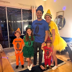 May include: A family of five wearing crayon-themed costumes. The group includes a blue crayon t-shirt, a yellow crayon t-shirt with a tulle skirt, a green crayon t-shirt with a tulle skirt, an orange crayon t-shirt with pants, and a red crayon t-shirt with pants.