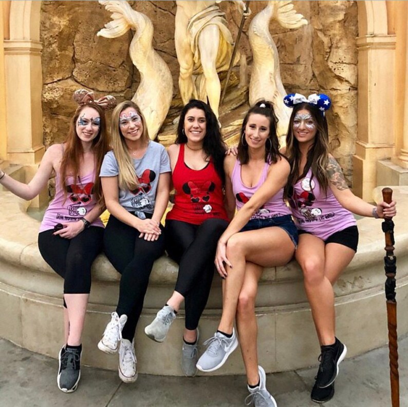 Peut inclure: Cinq personnes assises sur un rebord en pierre portent des v&ecirc;tements &agrave; th&egrave;me. Le groupe porte des d&eacute;bardeurs et des t-shirts avec un motif Mickey Mouse. Les chemises sont de diff&eacute;rentes couleurs, dont le rose, le rouge et le gris. Certains portent des shorts et des leggings.