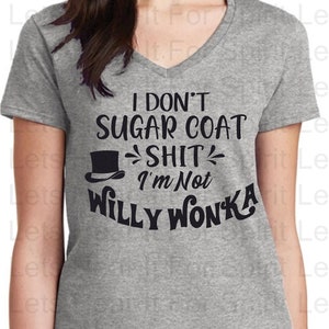 May include: A grey V-neck t-shirt with the text "I DON'T SUGAR COAT SHIT I'M NOT WILLY WONKA" in black. The shirt features a top hat graphic. The model is wearing blue jeans.