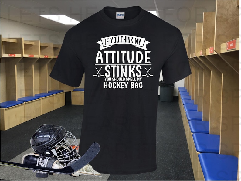 May include: A black t-shirt with white text that reads 'If you think my attitude stinks you should smell my hockey bag.' The t-shirt is displayed in a hockey locker room with hockey equipment on the floor.
