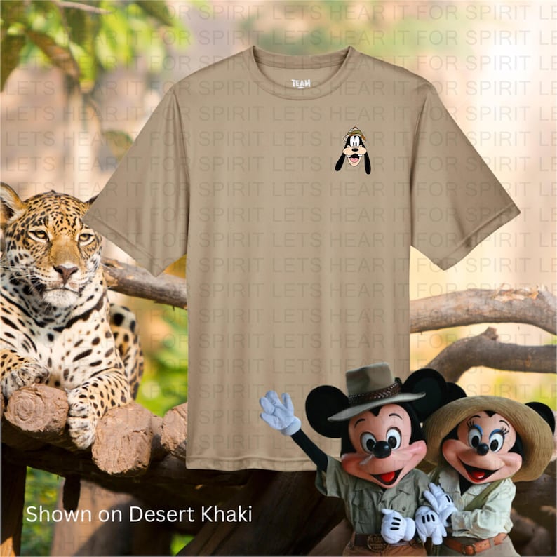 May include: Desert khaki t-shirt featuring a small embroidered image of Goofy wearing a safari hat. The shirt is displayed with a jaguar and costumed Mickey and Minnie Mouse characters. The text "Shown on Desert Khaki" is at the bottom.