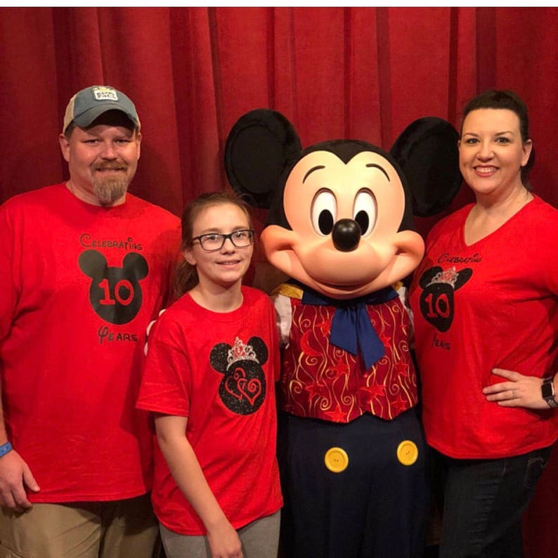May include: A family of three wearing red t-shirts with Mickey Mouse designs celebrating 10 years. The shirts feature a black Mickey Mouse head with the number 10 inside. The family is posing with Mickey Mouse in a red vest and blue pants.