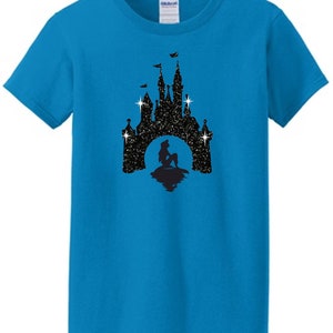Puede incluir: Una camiseta turquesa con una silueta negra de un castillo, una sirena y estrellas. El castillo tiene banderas y est&aacute; sobre un fondo estrellado. La camiseta est&aacute; hecha de un material suave.