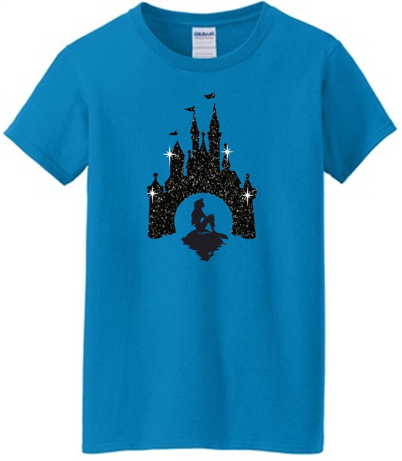 Puede incluir: Una camiseta turquesa con una silueta negra de un castillo, una sirena y estrellas. El castillo tiene banderas y est&aacute; sobre un fondo estrellado. La camiseta est&aacute; hecha de un material suave.