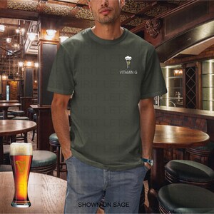 K&ouml;nnte beinhalten: Eine Person tr&auml;gt ein salbeigr&uuml;nes T-Shirt mit einer kleinen Grafik eines Biers und dem Text "VITAMIN G". Ein Pint Bier steht auf einem Holztisch in einem Pub.