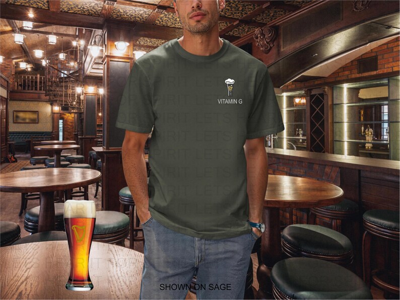 K&ouml;nnte beinhalten: Eine Person tr&auml;gt ein salbeigr&uuml;nes T-Shirt mit einer kleinen Grafik eines Biers und dem Text "VITAMIN G". Ein Pint Bier steht auf einem Holztisch in einem Pub.