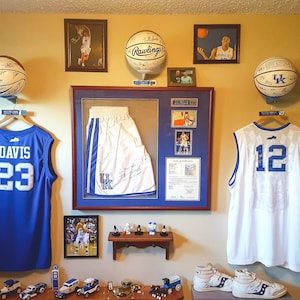 May include: A wall decorated with Kentucky Wildcats basketball memorabilia, including jerseys, basketballs, and framed photos. The jerseys are blue and white, with the numbers 21, 23, 12, and 00. The basketballs are signed and have the Kentucky Wildcats logo. The framed photos are of basketball players in action.