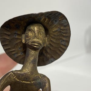 May include: A gold-colored metal figurine of a person wearing a wide-brimmed hat. The figurine has a detailed face and a simple body.