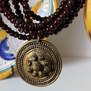 Pu&ograve; includere: Una collana multifilo realizzata con perline rosso scuro con un grande ciondolo rotondo in oro con un design decorativo.
