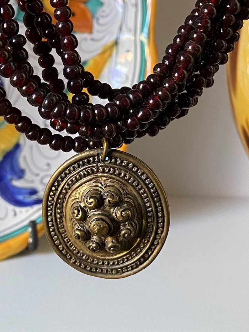 Pu&ograve; includere: Una collana multifilo realizzata con perline rosso scuro con un grande ciondolo rotondo in oro con un design decorativo.