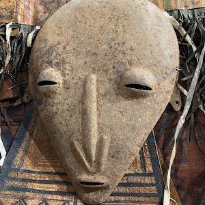 Puede incluir: Una máscara de metal envejecido, con una parte superior redondeada y una nariz larga y estrecha. La máscara tiene ojos pequeños en forma de hendidura y una boca rectangular. La superficie es de color marrón moteado, con signos de edad y desgaste. Es probable que sea un objeto decorativo o ceremonial.