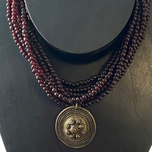 Pu&ograve; includere: Una collana multifilo realizzata con perline rosso scuro con un grande ciondolo rotondo, ornato e color oro.
