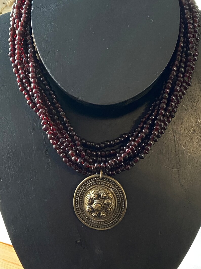 Pu&ograve; includere: Una collana multifilo realizzata con perline rosso scuro con un grande ciondolo rotondo, ornato e color oro.