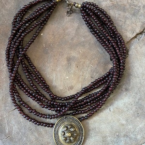 Pu&ograve; includere: Una collana multifilo realizzata con perline marroni scure con un grande ciondolo rotondo dorato con un design circolare.