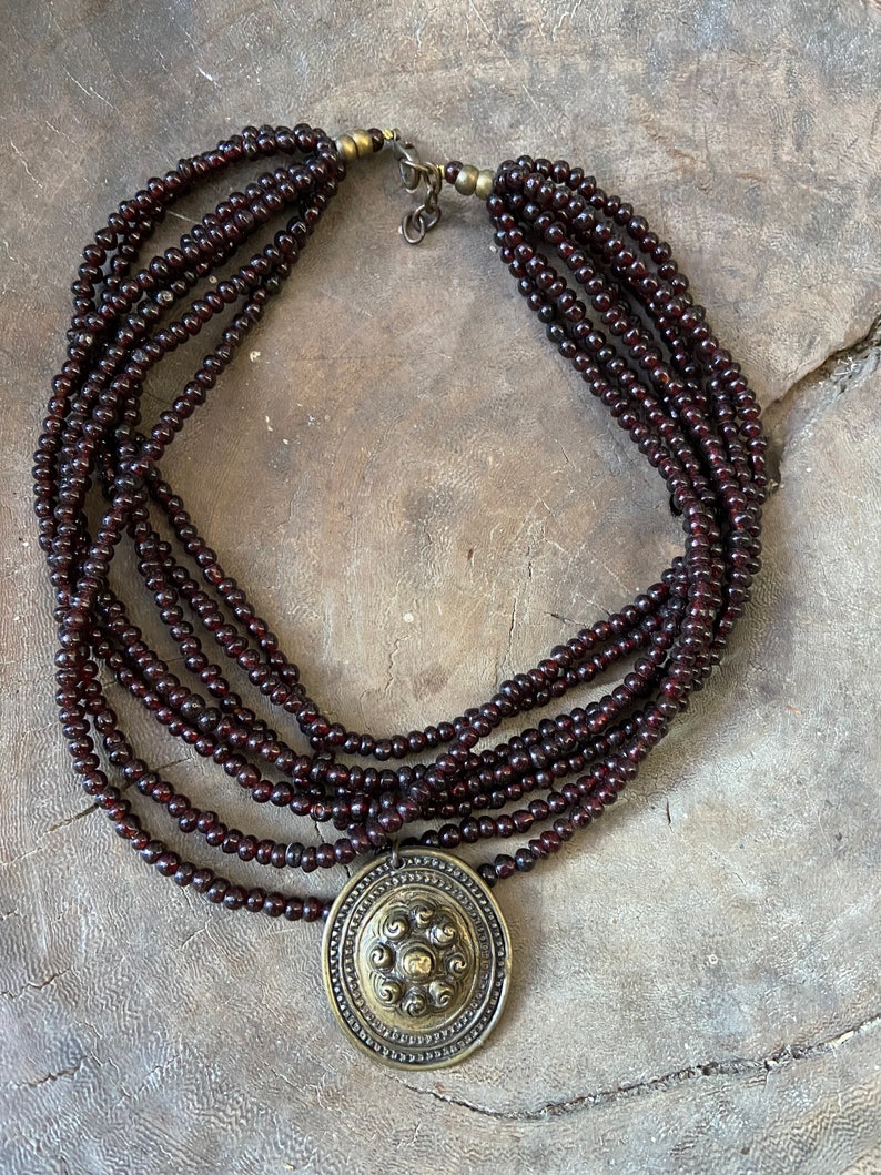Pu&ograve; includere: Una collana multifilo realizzata con perline marroni scure con un grande ciondolo rotondo dorato con un design circolare.