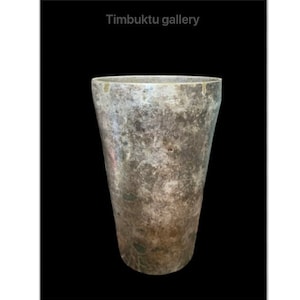 May include: A weathered, cylindrical vase with a wide mouth and a textured, mottled surface. The vase has a faded, aged appearance with shades of gray and brown. The text "Timbuktu gallery" is at the top.
