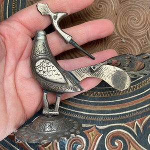 May include: A silver-coloured bird-shaped ornament with detailed patterns, possibly a decorative item or component. The bird features a detailed wing design and a separate, curved element. The background shows a wooden surface with carved patterns.
