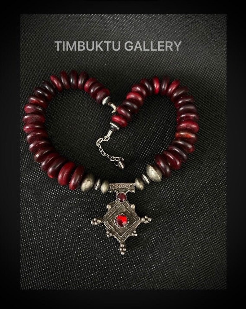 May include: A red wooden bead necklace with a silver pendant featuring a red gemstone. The pendant is in the shape of a cross with a decorative design. The necklace is displayed on a black background. TIMBUKTU GALLERY is written in white text at the top of the image.