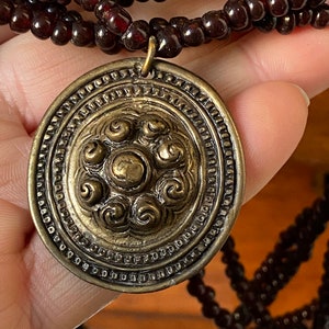 Pu&ograve; includere: Una collana multifilo realizzata con perline rosso scuro con un grande ciondolo circolare decorato. Il ciondolo &egrave; in ottone e presenta un design dettagliato di motivi a spirale.