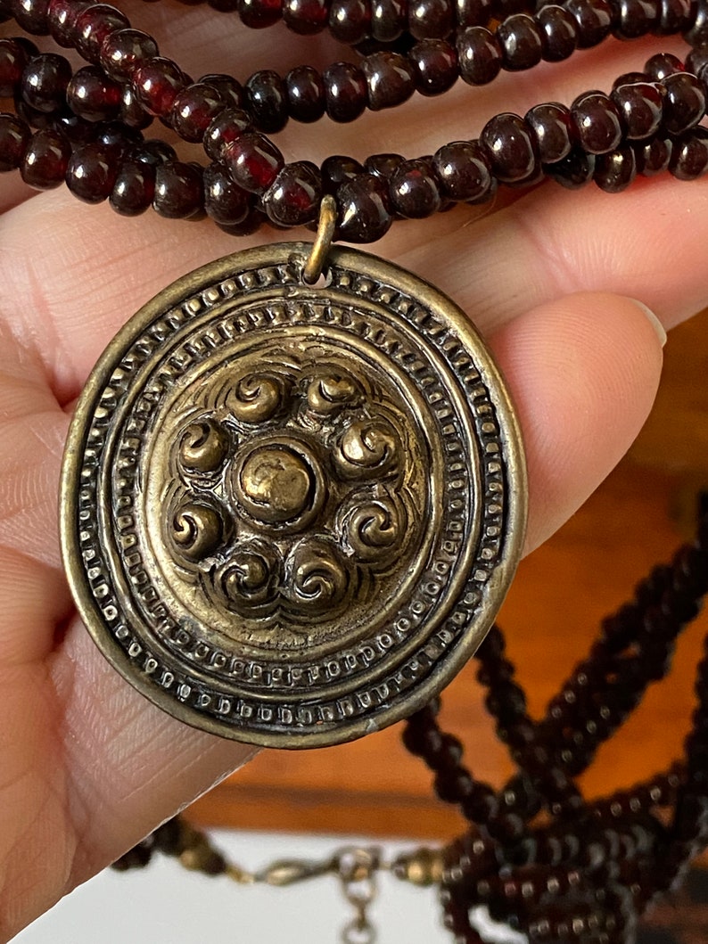 Pu&ograve; includere: Una collana multifilo realizzata con perline rosso scuro con un grande ciondolo circolare decorato. Il ciondolo &egrave; in ottone e presenta un design dettagliato di motivi a spirale.