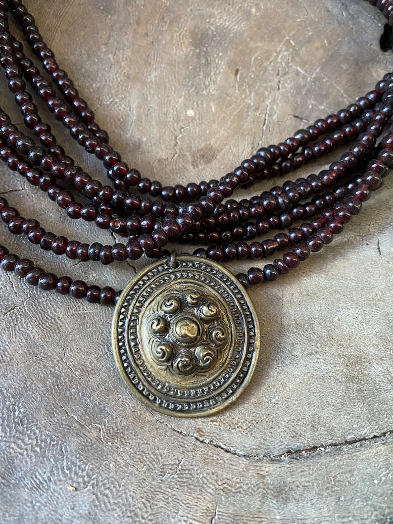 Pu&ograve; includere: Una collana di perline marroni scure con un grande ciondolo rotondo in ottone che presenta un design circolare con cinque vortici.