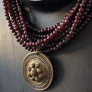 Pu&ograve; includere: Una collana multifilo realizzata con perline rosso scuro con un grande ciondolo rotondo color oro con un design decorativo.