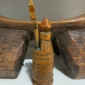 May include: A wooden bottle stopper with a carved design and a small wooden stick. The bottle stopper is brown and has a pointed top.
