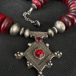 May include: A red glass bead necklace with a silver pendant featuring a cross design and red gemstones.