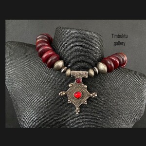 May include: A silver pendant necklace with a red stone and a geometric design. The necklace is made of dark red beads and is on a mannequin.