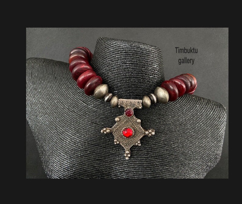 May include: A silver pendant necklace with a red stone and a geometric design. The necklace is made of dark red beads and is on a mannequin.