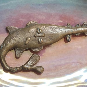 May include: A bronze-colored fish-shaped brooch. The brooch features a stylized design with fins, a tail, and a detailed body. The fish has a pin on the back for attachment. The brooch is likely vintage or antique.