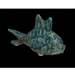 Può includere: Una statuetta di pesce in ceramica blu-verde con una superficie testurizzata. Il pesce ha una grande pinna dorsale e una bocca larga.