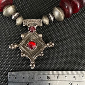 May include: A silver necklace with a red glass pendant and red wooden beads. The pendant is a square with a smaller square in the center. The necklace is measured with a ruler, showing it is 5 inches long.
