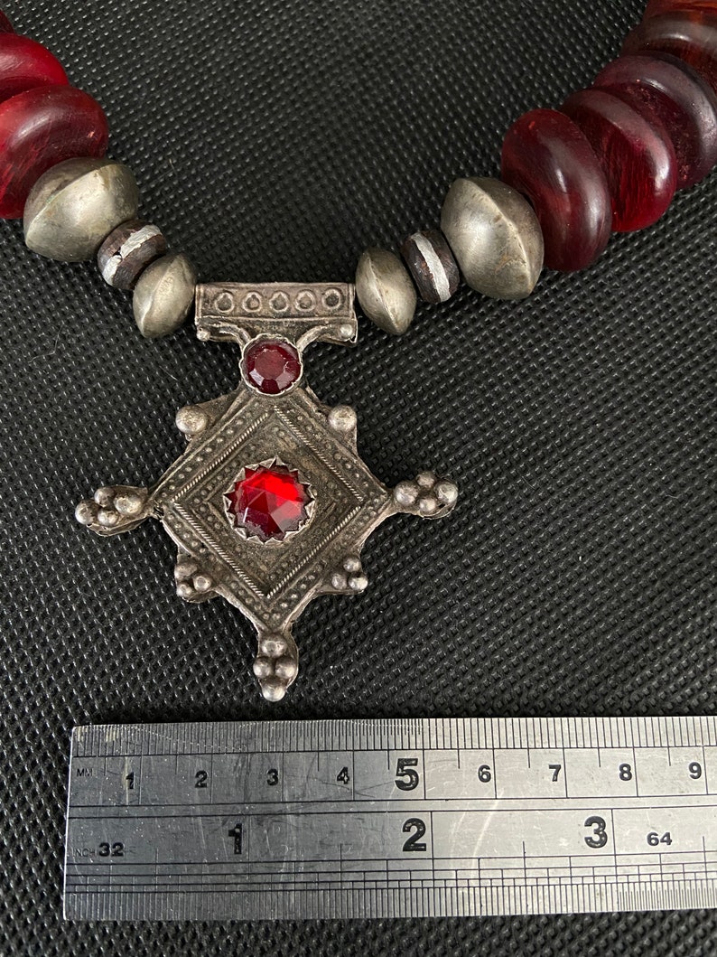 May include: A silver necklace with a red glass pendant and red wooden beads. The pendant is a square with a smaller square in the center. The necklace is measured with a ruler, showing it is 5 inches long.