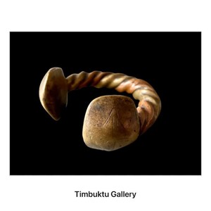 May include: A bronze bracelet with a twisted band and a square, engraved centerpiece. The bracelet has a weathered appearance, suggesting age and history. The background is black.