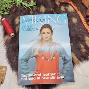 Peut inclure: Un magazine intitulé "VIKING DRESS CODE" avec Kamil Rabiega en couverture. L'image de couverture montre une femme vêtue d'une tenue de style scandinave. Le texte "Textile and leather clothing in Scandinavia" est également visible.