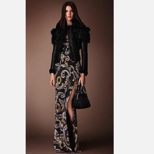 Puede incluir: Una mujer con una chaqueta de cuero negro con ribete de piel sobre un vestido largo negro con un estampado floral dorado y blanco. Sostiene un pequeño bolso negro.