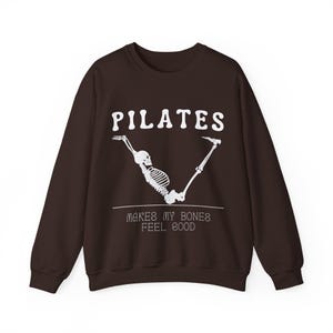 Puede incluir: Una sudadera marrón con el texto "PILATES makes my bones feel good" y un esqueleto blanco haciendo una pose de pilates.