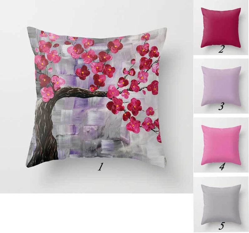 May include: A throw pillow with a pink and gray floral design. The pillow features a stylized painting of a tree with pink blossoms. The pillow is shown against a white background.