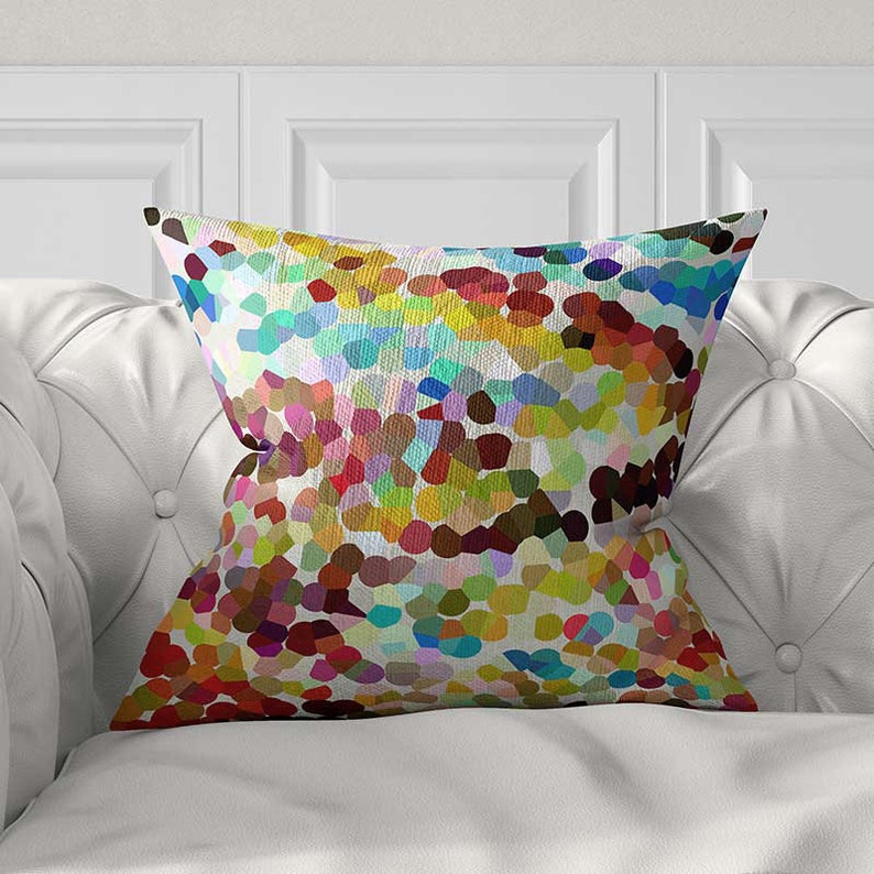 May include: A square throw pillow featuring a vibrant, multi-colored mosaic pattern composed of small, irregular shapes in shades of red, yellow, blue, green, and purple. The fabric has a visible woven texture, creating a pointillist effect.